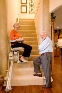 senior couple looking at camera while woman is seated on indoor straight stair lift