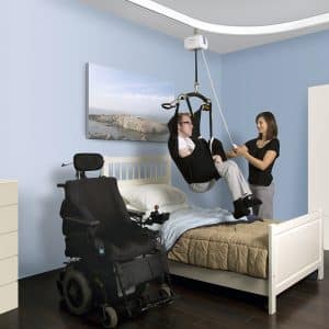 man using ceiling lift helped by caregiver with wheelchair next to the bed